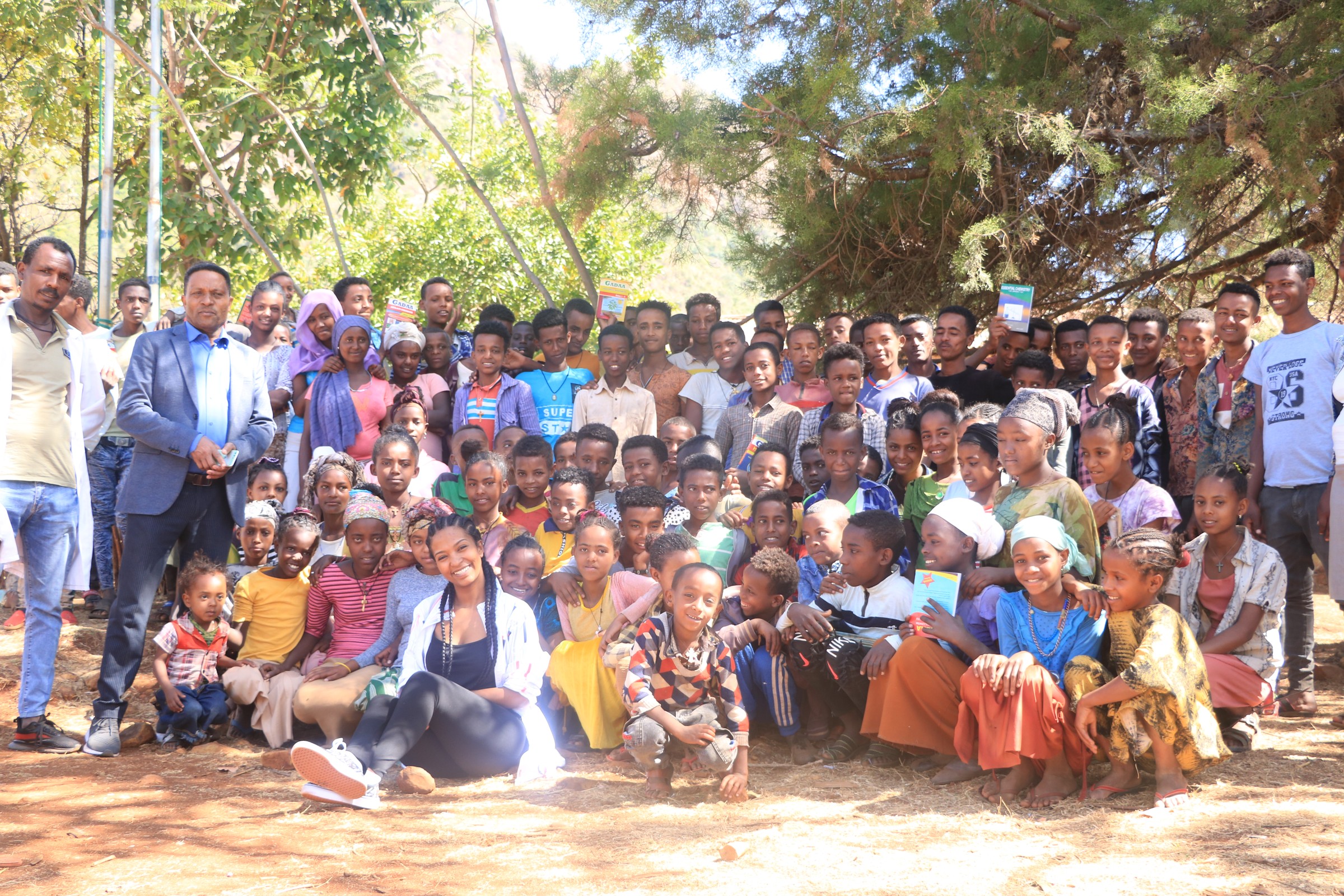 Adults and a large group of students gathered outdoors under trees, several holding books