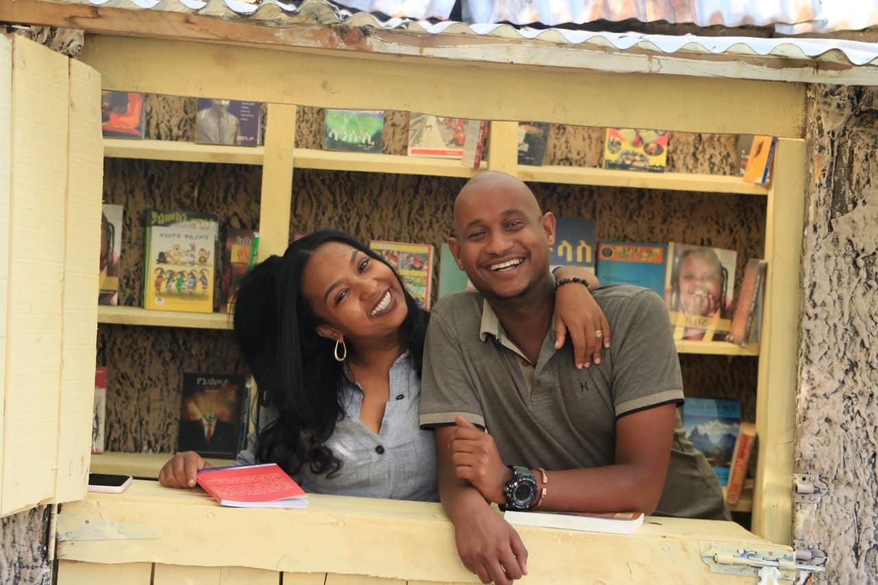 Two volunteers smiling at the window of the Sinen book club kiosk, shelves of books behind them